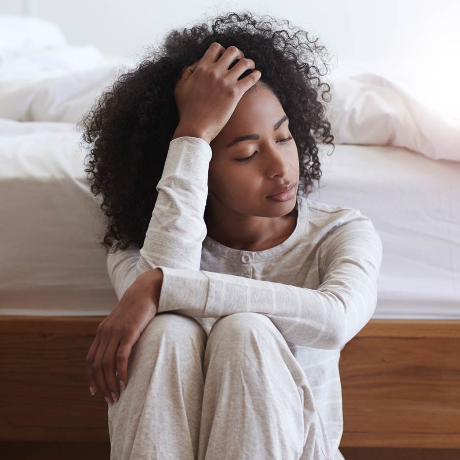 Young black woman sitting against bed with headache